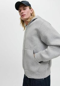 Grå zip-up hættejakke lavet af blødt stof, med en frontlomme, justerbar hætte og ribbede manchetter, parret med mørk denim.