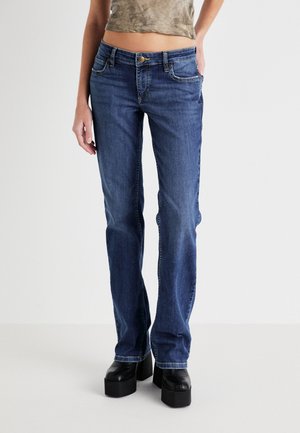 Vrouw met mid-rise, rechtvallende blauwe spijkerbroek, zwarte plateauzolen en een cropped groen topje, staand tegen een effen lichte achtergrond.