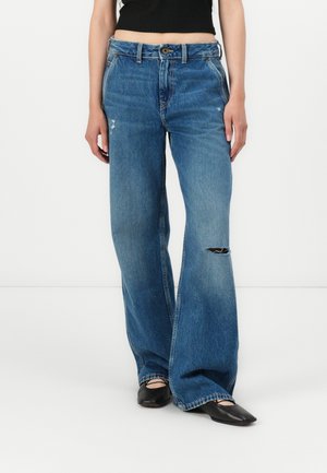 Femme portant un jean bleu taille haute à jambes larges avec une déchirure au genou, un top court noir et des ballerines noires pointues sur fond blanc.