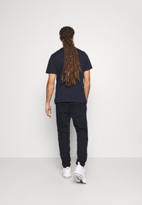 Navyfarbens Baumwoll-T-Shirt und schwarze Jogginghosen, getragen mit weißen Sneakers. Langes Haar nach hinten gebunden, von hinten betrachtet vor einem schlichten Hintergrund.