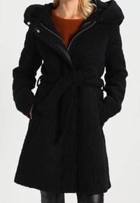 Mustat, tekstuurilla varustetut takki, jossa on huppu, vyötäröllä vyö ja vetoketjulla suljettava etuosa. Sisältää sivutaskut ja pehmeän, pörröisen kankaan.