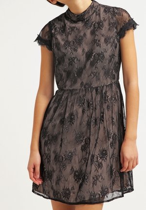 Frau trägt ein kurzes schwarzes Blumenspitzenkleid mit transparenten kurzen Ärmeln und hohem Kragen, steht vor einem einfachen Hintergrund.