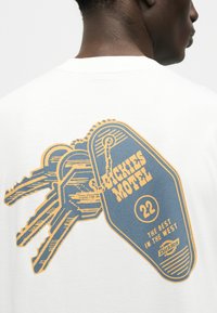 Άνδρας που φοράει λευκό πουκάμισο με γραφικό μιας μπρελόκ μοτέλ με την ετικέτα "Dickies Motel 22 The Best in the West" και τρία προσαρτημένα κλειδιά.