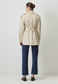 Camisa beige de gran tamaño con cinturón en la cintura, mangas largas y hombreras, combinada con jeans de pierna recta azul oscuro y zapatos marrones.