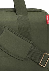 Borsa di tessuto verde oliva con superficie texture, dotata di una larga tracolla fissata da una fibbia in metallo dorato e un'etichetta logo rossa sul lato.