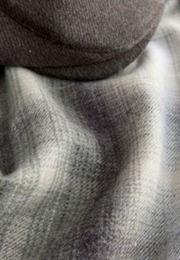 Woolen blanket in muted tones of gray and beige, featuring a checked pattern, soft texture, and a rounded brown wool cap partially visible.