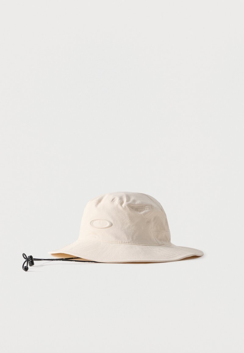 Chapeau bob beige à large bord avec sangle noire ajustable sous le menton, posé à plat sur un fond neutre.