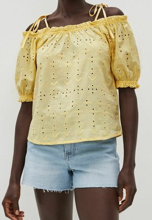 Top giallo off-the-shoulder con maniche a sbuffo, caratterizzato da motivi floreali ritagliati e scollatura arricciata. Abbinato a pantaloni denim.