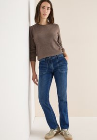 Jean en denim bleu avec une coupe décontractée, dotée de poches avant et d'une taille réglable par cordon, associée à un pull côtelé marron et des baskets beiges.