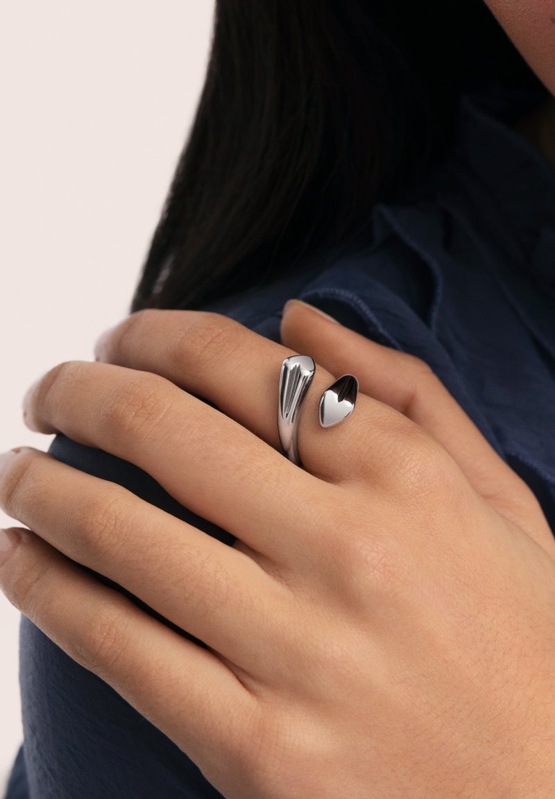 Mano con anillo ajustable plateado con un corazón y un diseño abstracto que descansa sobre el hombro, con cabello oscuro y ropa azul marino visibles.