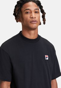 Svart bomull t-shirt med ribbat krage, korta ärmar och en liten broderad logotyp i rött, vitt och blått på bröstet.