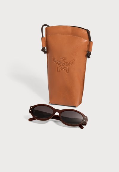 Lunettes de soleil ovales marron avec verres foncés à côté d'une grande pochette en cuir fauve embossée du logo MCM et fermeture à cordon.
