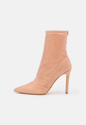 Brzoskwiniowy botek na wysokim obcasie, pokryty małymi, błyszczącymi kryształkami, z zaostrzonym noskiem i smukłym obcasem typu szpilka.