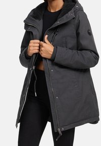 Femme portant un manteau d'hiver noir à zip et capuche avec cordons, en train de fermer la fermeture éclair, accompagné d'un pantalon et d'un pull noir.