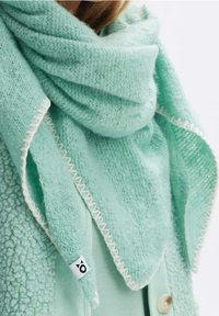 Écharpe vert menthe clair en tissu doux et texturé. Présente des coutures blanches le long des bords et une petite étiquette de marque dans un coin.