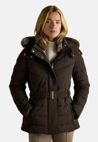 Tmavo hnedý vypchávaný kabát s kapucňou s kožušinovým lemovaním, so zipsom a gombíkovým zapínaním, nastaviteľným pásom a prešívaným vzorom. Nosený cez biely sveter.