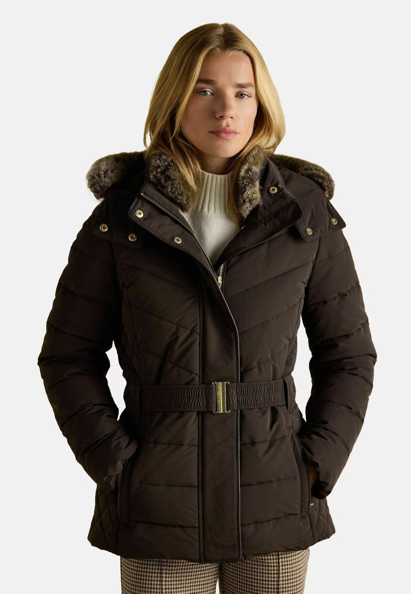 Tmavo hnedý vypchávaný kabát s kapucňou s kožušinovým lemovaním, so zipsom a gombíkovým zapínaním, nastaviteľným pásom a prešívaným vzorom. Nosený cez biely sveter.