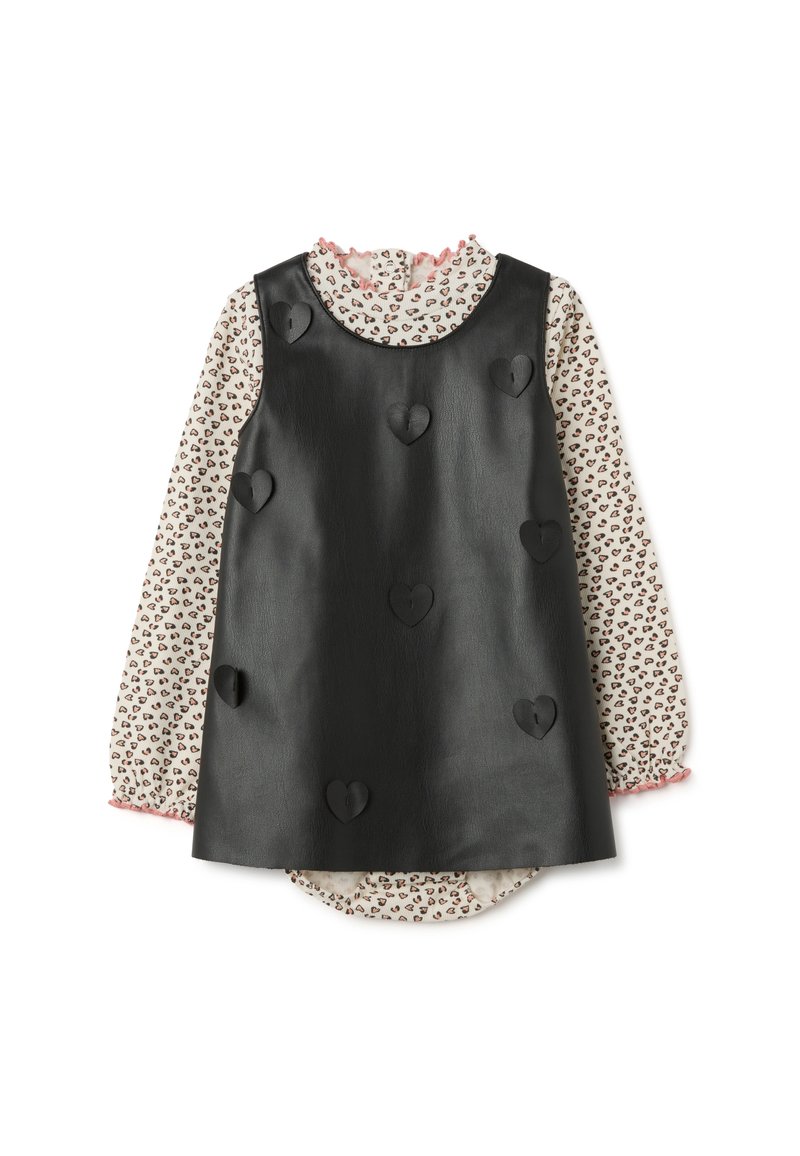 Schwarzes Kunstleder-Kleid ohne Ärmel mit gestickten Herzdetails über einer langärmligen cremefarbenen Bluse mit kleinen Herzmotiven und pinken Rüschensäumen.