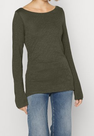 Vrouw draagt een olijfgroen shirt met lange mouwen, gecombineerd met verweerde blauwe jeans, staand tegen een effen lichte achtergrond.