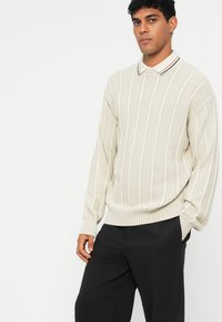 Pulôver de malha em creme com riscas verticais, apresentando um pequeno colarinho com riscas pretas e brancas. Estilizado com calças pretas.