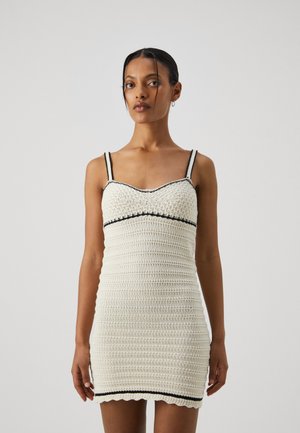 White knitted dress with thin black straps, featuring a scalloped hem and textured pattern. The design is form-fitting, highlighting the silhouette.