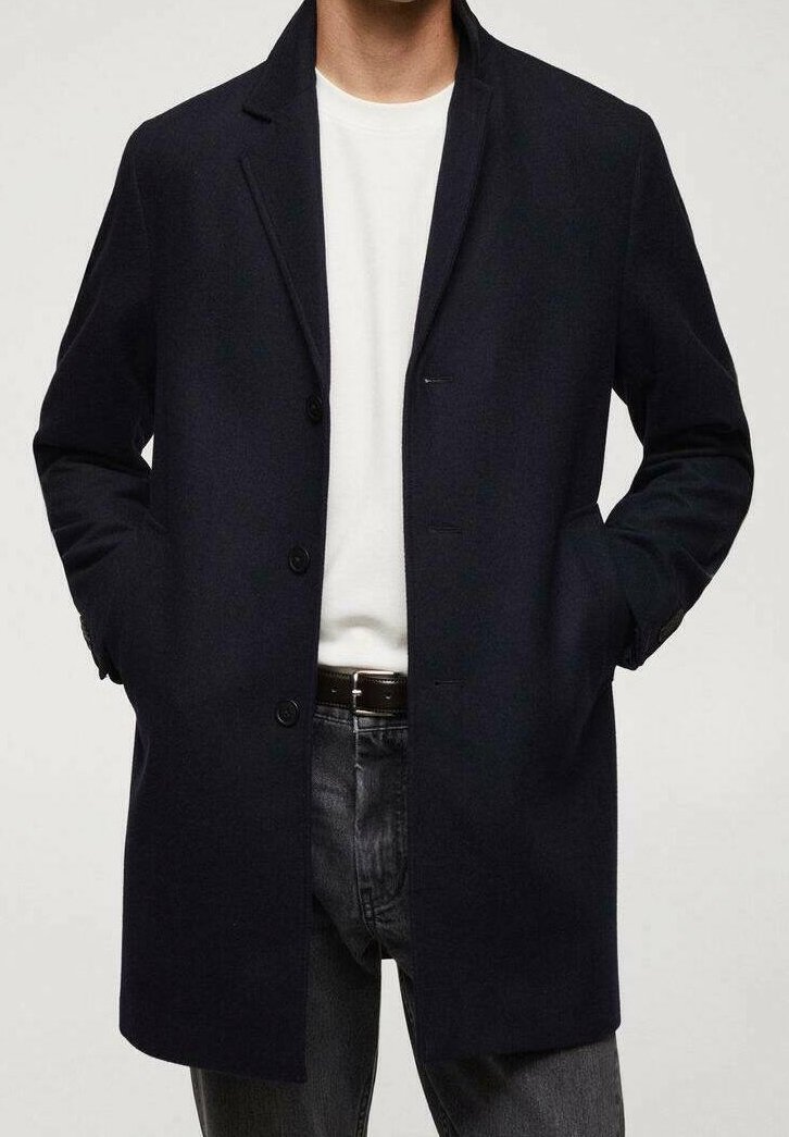 Homme portant un manteau bleu marine foncé par-dessus une chemise blanche et un jean gris, les mains dans les poches du manteau, debout devant un fond neutre.