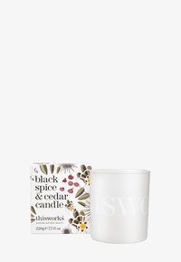 Weiße zylindrische Kerze mit dem eingeprägten Schriftzug "thisworks". Begleitet von einer dekorativen Schachtel mit floralen Mustern und dem Text "black spice & cedar".
