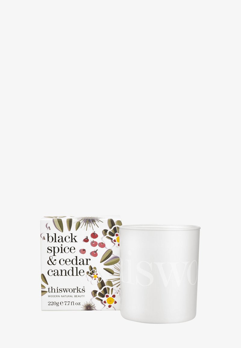 Weiße zylindrische Kerze mit dem eingeprägten Schriftzug "thisworks". Begleitet von einer dekorativen Schachtel mit floralen Mustern und dem Text "black spice & cedar".