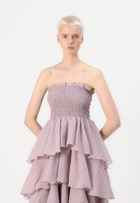 Strapless kjole i blød lavendelfarvet stof med en smocket bodice og lagdelt volangnederdel. Let tekstur og svajende silhuet.