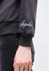 Άτομο που φοράει σκουρόχρωμα τζιν και σκουρόχρωμο φούτερ με κεντημένο το "Magnolia" στο μανίκι, χέρι χαλαρά στο πλάι με ένα δαχτυλίδι.