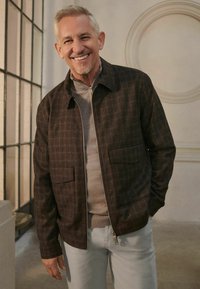Brown plaid zip-up jacket with two front pockets, worn over a light gray sweater and light gray pants. Smooth texture and tailored fit.