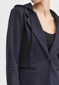 Blazer blu navy con cappuccio, realizzato in tessuto morbido. Presenta una chiusura con un bottone e una tasca laterale, con una silhouette aderente.