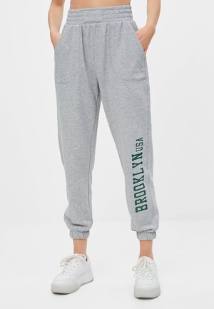 Osoba nosząca jasnoszare dresy z elastycznymi mankietami przy kostkach i nadrukiem „BROOKLYN USA” pionowo na lewej nodze, w połączeniu z białymi trampkami.