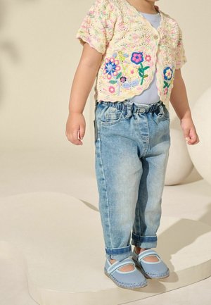 Tout-petit portant un jean bleu clair, des chaussures Mary Jane bleues et un cardigan court en tricot crème avec une broderie florale colorée.