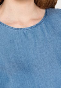 Primo piano del collo e della parte superiore del torace di una persona che indossa una camicia di denim azzurro chiaro con scollatura rotonda.