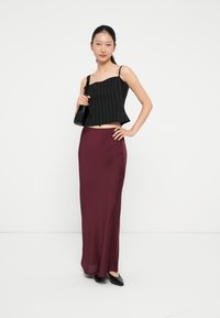 Fekete csíkozott crop top pántos vállpántokkal, burgundi bordó, földig érő szoknyával párosítva. A modellen fekete lapos cipők vannak, fehér háttér előtt áll.