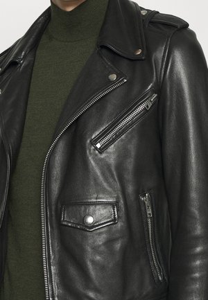 Schwarze Leder-Bikerjacke mit silbernen Reißverschlüssen und Druckknöpfen, getragen über einem olivgrünen Rollkragenpullover, Nahaufnahme.