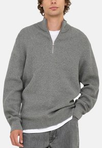 Grå hættetrøje med lynlås og ribbet tekstur samt høj krave. Har lange ærmer og en afslappet pasform. Bæres over en hvid t-shirt og grå jeans.
