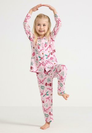 Vaaleanpunaiset puuvillapijamat, joissa on kukka- ja nauhatulostuksia, sisältävät napitettavan yläosan ja yhteensopivat housut. Lapsi poseeraa leikkisästi, paljain jaloin.