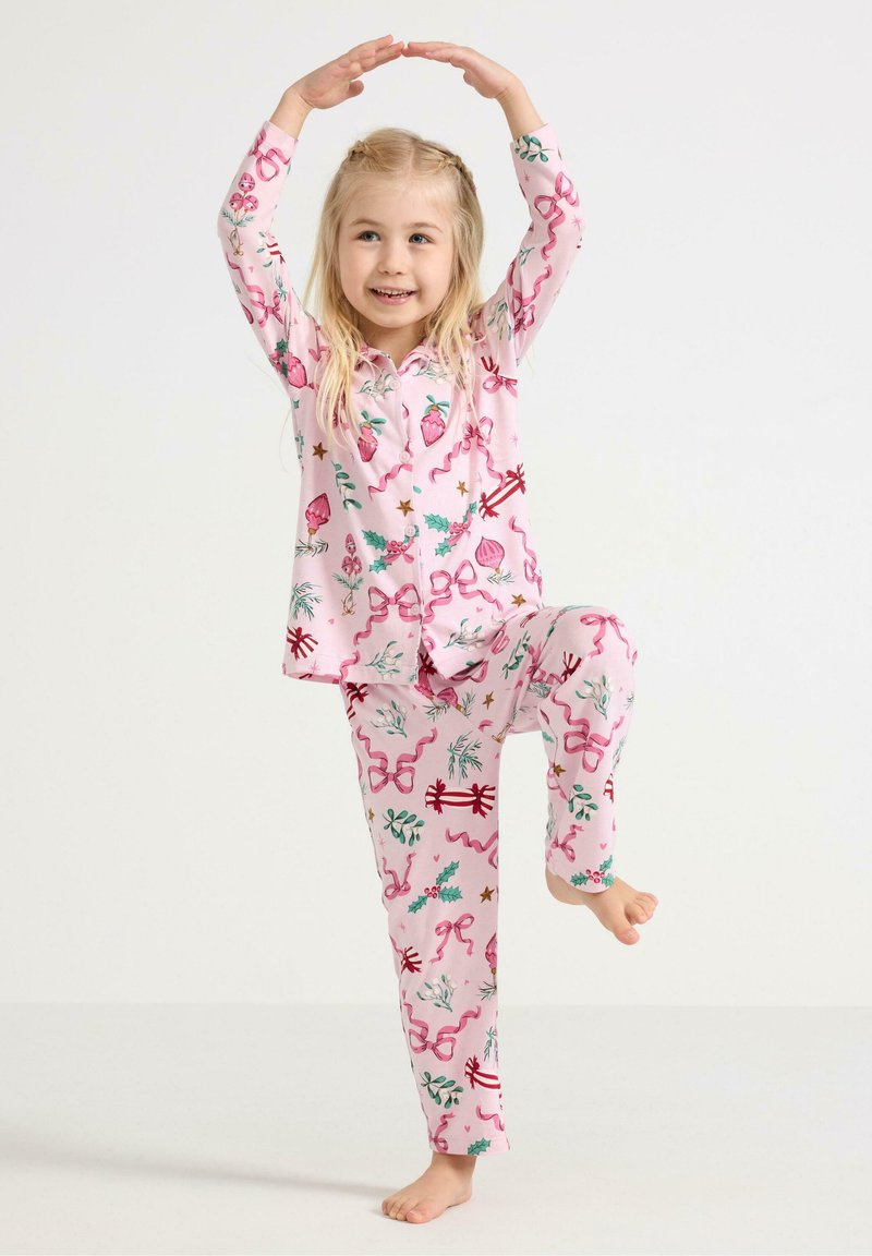 Rosa Baumwollschlafanzüge mit Blumen- und Bandmustern, bestehend aus einem knöpfbaren Oberteil und passenden Hosen. Das Kind posiert spielerisch, barfuß.