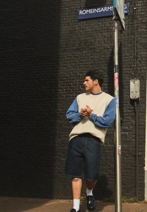 Mand i blå langærmet skjorte og beige vest læner sig op ad en stolpe under et gadeskilt med teksten "Romeinsarms" mod en sort murstensvæg.