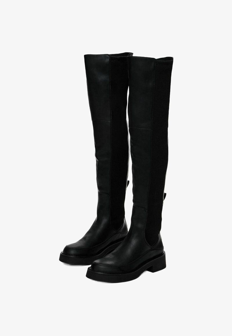 Bottes noires en cuir montantes jusqu'aux genoux avec des panneaux latéraux élastiques et des talons bas épais, disposées en biais côte à côte sur un fond blanc.