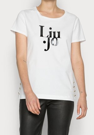 Mujer que lleva una camiseta blanca con texto negro "Liu Jo" y pantalones de cuero negros, de pie con los brazos relajados a los lados.