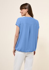 Blusa azzurro chiaro con maniche corte arrotolate, orlo arricciato e uno sbieco sul retro. Il materiale appare liscio con una vestibilità rilassata. Abbinata a pantaloni bianchi.