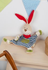 Soft bunny toy with red ears and scarf, blue and white striped body with green clover, sitting on a light wooden desk under colorful bunting.