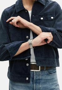 Marinblå kordjacka med knäppning framtill, två bröstfickor och uppvikta ärmar; kombinerad med en vit skjorta och denimjeans.