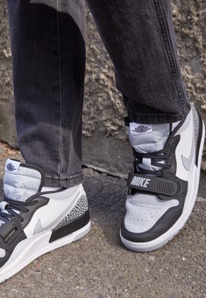 Person wearing black and white Nike Air Jordan sneakers standing on a rough pavement near a textured stone wall.