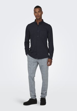 Camisa de manga larga de color azul marino con botones en la parte delantera y un bolsillo en el pecho, combinada con pantalones ligeros de cuadros grises y zapatos negros.