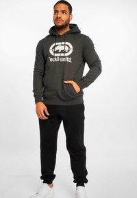 Haut à capuche gris foncé avec une poche avant et un cordon de serrage, arborant un logo graphique blanc. Assorti à un pantalon de survêtement noir et des baskets blanches.