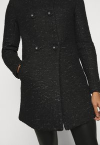 Dubbelknäppt svart kappan i texturerat tyg, med stora svarta knappar och sidofickor, samt en något avsmalnande silhuett.